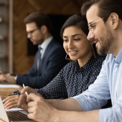 In modern office diverse colleagues Indian businesswoman and caucasian businessman workmates talking sit at desk coworking space use laptop, discuss work result, new business professional application